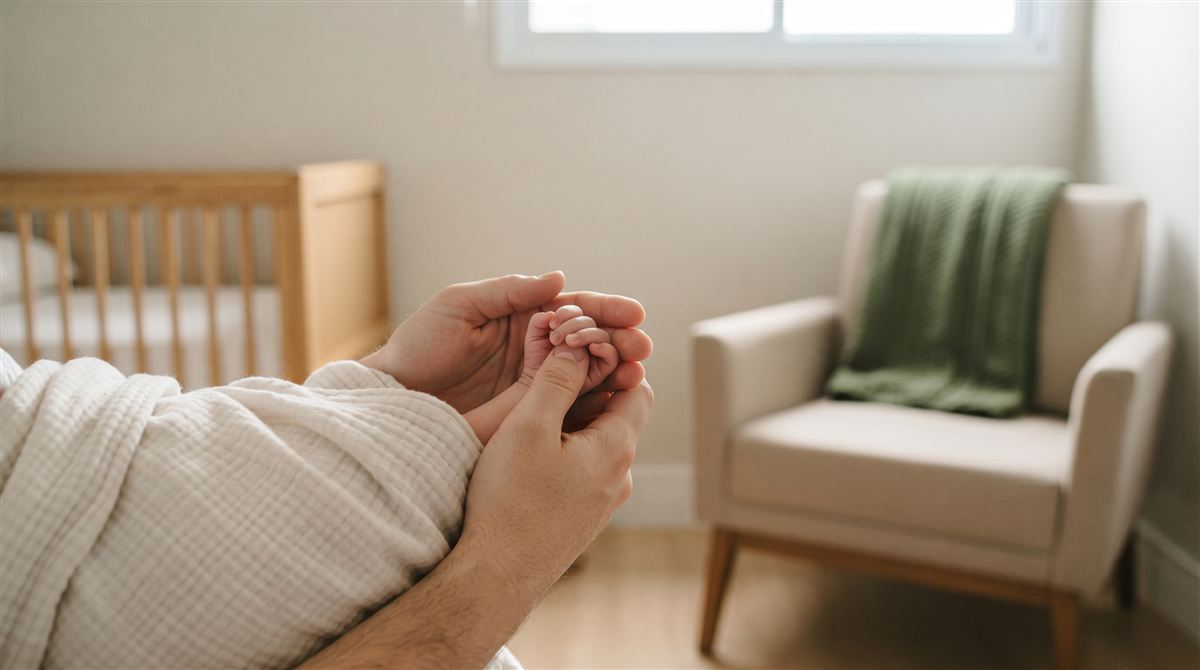 Mãos de um adulto envolvendo a mão de um recém-nascido, com um berço ao fundo: licença-maternidade e paternidade do servidor público